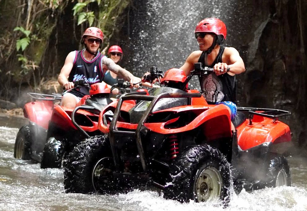 ATV riders splashing through a river by a waterfall in Bali